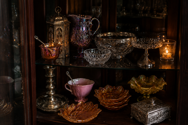 A collection of vintage glassware and decorative tableware displayed on dark wooden shelves, including a pink teacup, iridescent glass pitchers, cut crystal bowls, amber glass plates, and silver-toned metal containers.