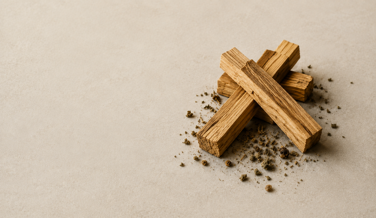 Three small wooden incense sticks arranged in a cross shape on a light beige textured surface, with scattered dark incense powder around them.