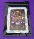 A black holographic pouch containing a 'Retrograde Reset Cord Cutting Ritual Kit' with an illustrated black label, placed on a purple background.