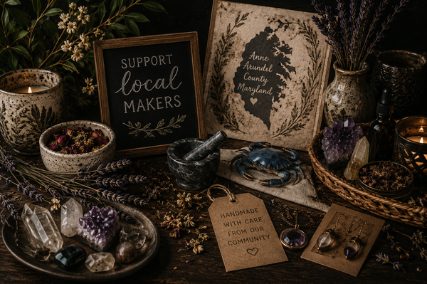 A styled tabletop arrangement in a dark rustic setting featuring handmade jewelry, crystals, dried lavender, candles, a small chalkboard sign reading 'SUPPORT local MAKERS,' a framed map-style sign, and a handmade tag that says 'HANDMADE WITH CARE FROM OUR COMMUNITY.'.